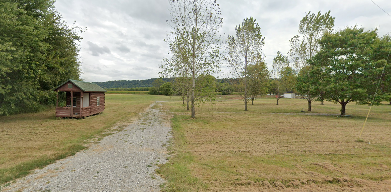 A small wooden cabin sits beside a gravel path in an open field with scattered trees under a cloudy sky.