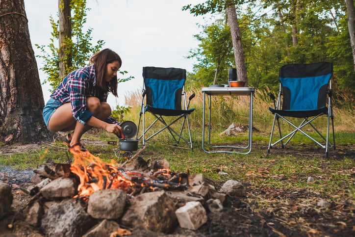 A woman crouches by a campfire, pouring water from a kettle. Two chairs and a table with a cup are nearby. Trees and grass surround the campsite.