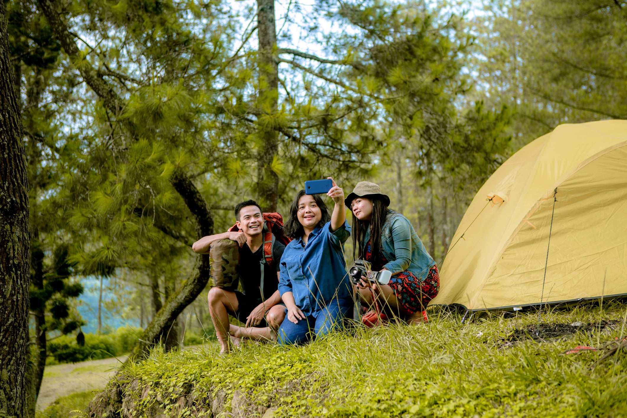 Three people taking a selfie in front of a yellow tent in a forested area, surrounded by greenery and trees.