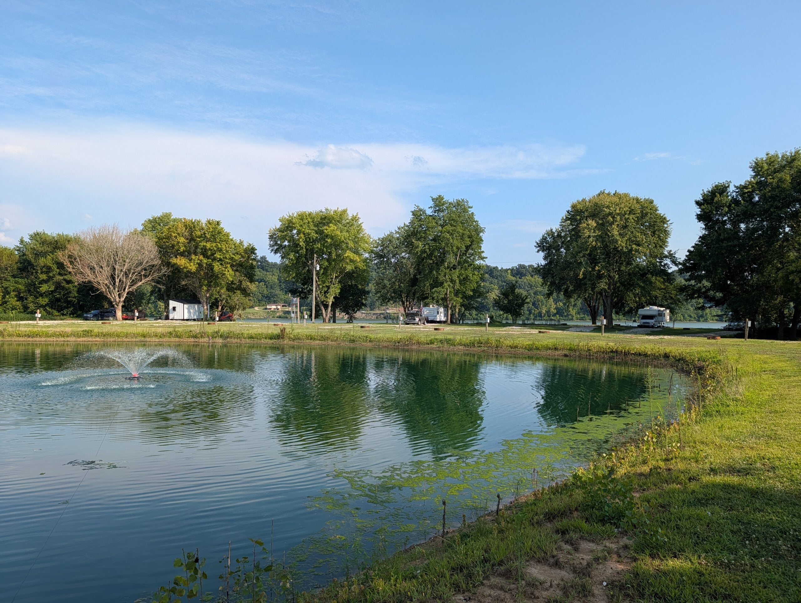 A small pond with a water fountain is surrounded by grass and trees, with a few trailers and vehicles visible in the background under a clear blue sky.