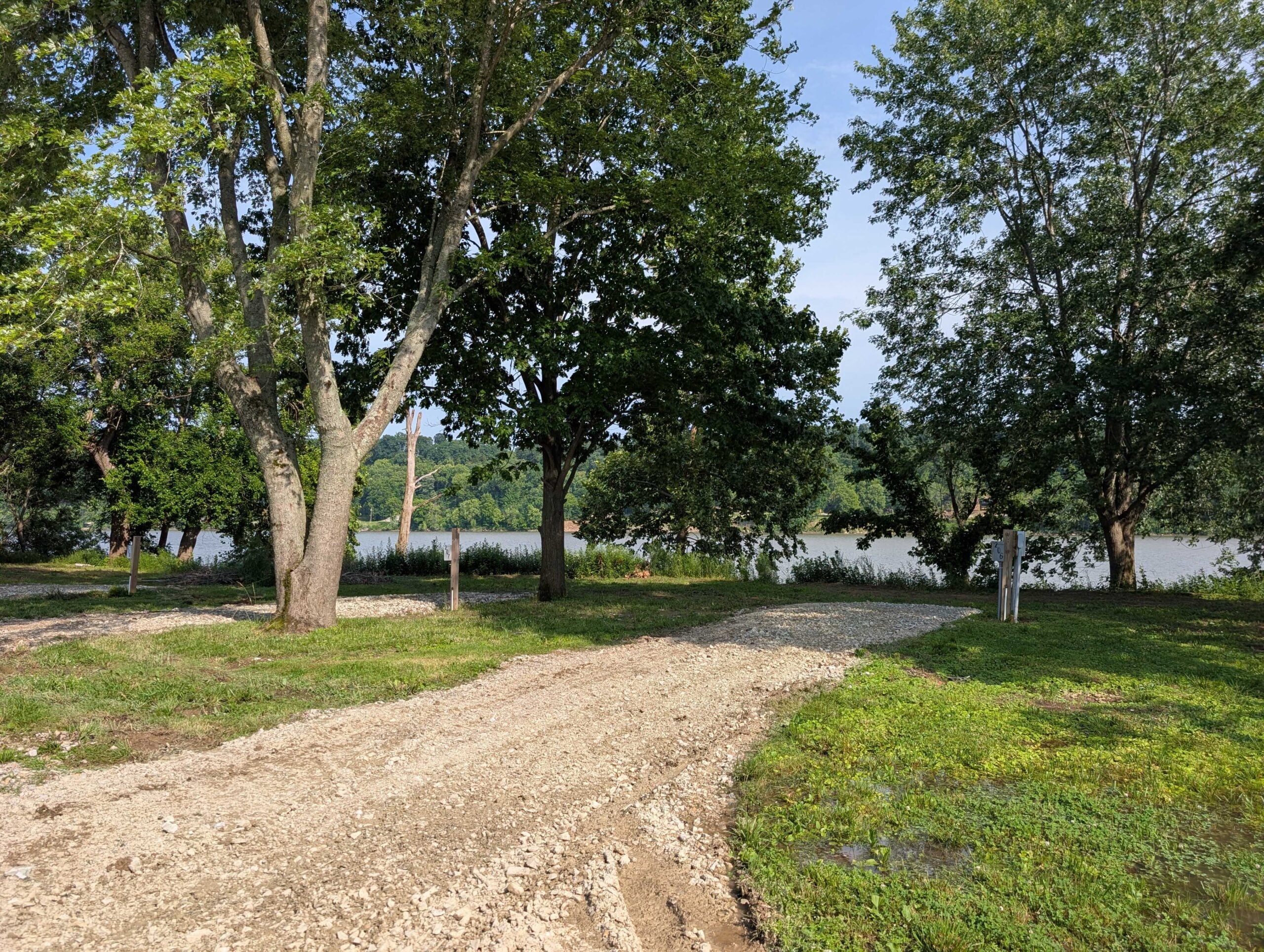 Gravel path leads through green grass and trees toward a calm river under a clear blue sky.