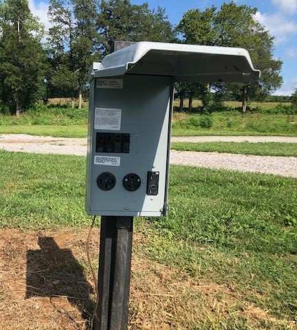 A gray outdoor electrical panel with a cover is mounted on a black post in a grassy area, near a gravel path and trees.